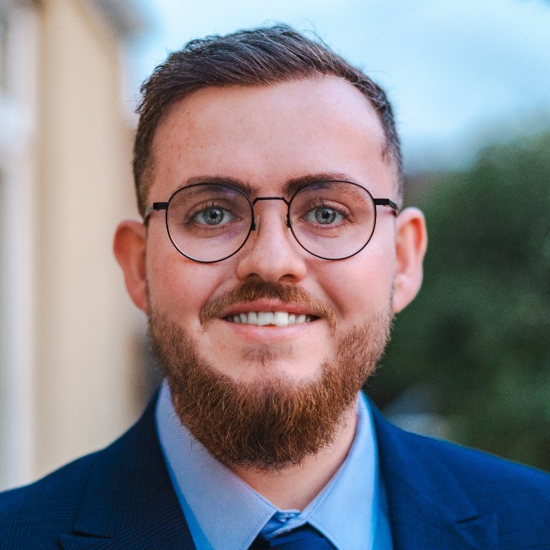 Portrait of a smiling man with a beard and glasses, wearing a suit and tie. he stands outdoors with a blurred building background.