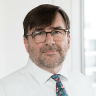 A middle-aged man wearing glasses, a white shirt, and a colorful tie, smiling subtly in a bright, office-like setting.