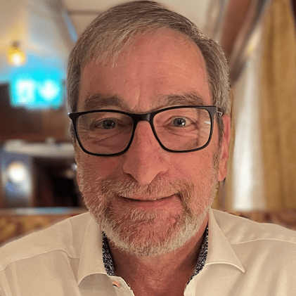 A man with glasses, light beard, and short grey hair wearing a white shirt is looking at the camera. The background is a softly lit indoor setting. - SysAid