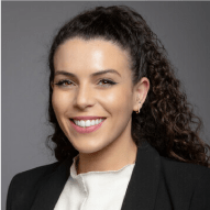 Professional headshot of a smiling woman with curly hair, wearing a black blazer and white top, against a gray background.