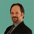 A man with a beard and mustache smiling, wearing a dark suit and tie, standing against a teal background.