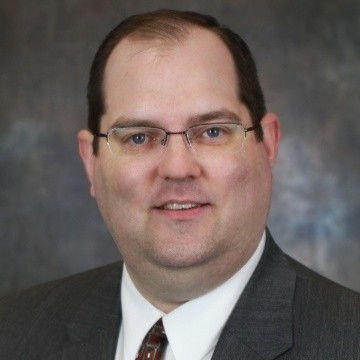 Portrait of a middle-aged man wearing glasses, a white shirt, and a striped tie with a gray suit jacket, smiling against a gray background.