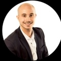 A man with a shaved head, wearing a black blazer and a white shirt, smiling against a white background.