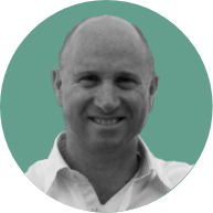 Black and white photo of a smiling bald man in a white shirt, set against a plain, teal background.