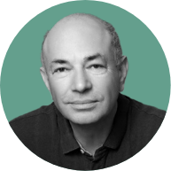 Portrait of a middle-aged bald man with a gentle smile, wearing a dark shirt against a teal background.