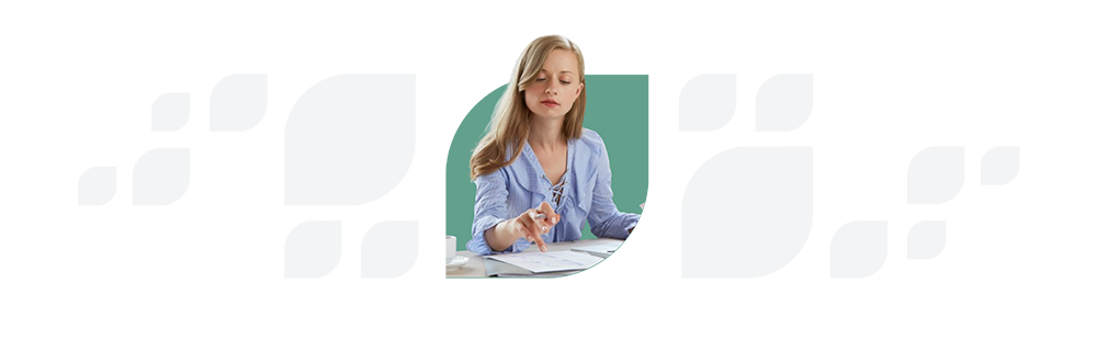 A woman in a blue shirt is focused on writing notes at a desk surrounded by papers.