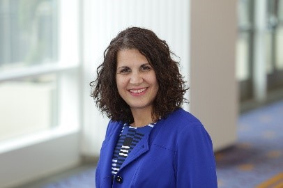 A professional woman with curly hair, wearing a blue blazer, smiling in a brightly lit indoor space.